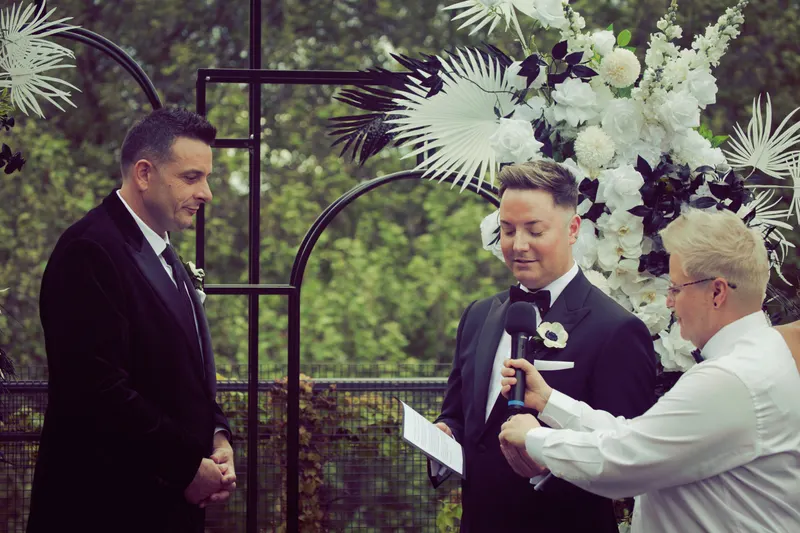 A groom speaks his vows into a microphone held by Lizanne, beside a floral arch