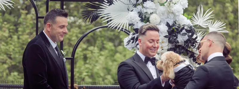 A groom in a bow tie smiles down at a small dog during the ceremony